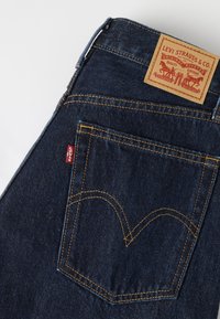 Jean Levi's bleu foncé avec poche arrière visible, couture orange, étiquette rouge Levi's et patch en cuir marron sur la ceinture.