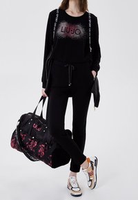 Schwarzes Sweatoutfit mit glitzerndem "LIU-JO"-Logo, ausgestattet mit einer schwarzen Reisetasche mit floralen Akzenten und mehrfarbigen sportlichen Turnschuhen.