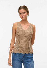 Top de punto beige sin mangas con escote en V, que presenta un patrón en forma de chevrón y un dobladillo festoneado, combinado con jeans azules. Tela texturizada y diseño sin mangas.