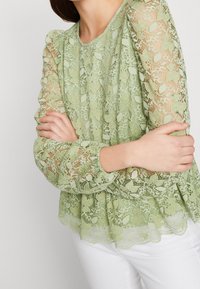 Donna con le braccia incrociate, che indossa una blusa a maniche lunghe in pizzo floreale verde chiaro e pantaloni bianchi, vista dal collo ai fianchi.