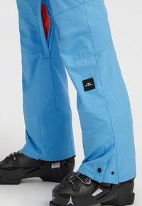 Blauwe skibroek gemaakt van waterproof stof met verstelbare manchetten, voorzien van een zwart logo patch en ritssluitingen aan de zijkant voor ventilatie.