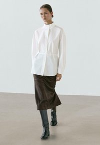 Wit shirt met lange mouwen, een hoge kraag en knopen. Gecombineerd met een donkerbruine rok van middellange lengte en strakke zwarte kniehoge laarzen.