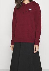 Bordeaux hoodie med framficka, långa ärmar, dragsko i huvan och vit Nike-logotyp på bröstet, matchad med en svart kjol.