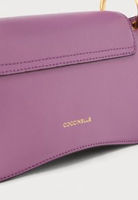 Μωβ δερμάτινη τσάντα χειρός με χρυστό κρίκο-χερούλι και χρυστό λογότυπο COCCINELLE που είναι ανάγλυφο στο μπροστινό καπάκι με εμφανείς ραφές.