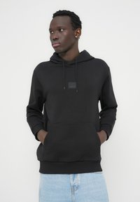 Μαύρο φούτερ με κουκούλα, μπροστινή τσέπη, στενές μανσέτες και μικρό λογότυπο σε ύφασμα. Φοριέται με ανοιχτόχρωμο τζιν. Ομαλή υφή υφάσματος.