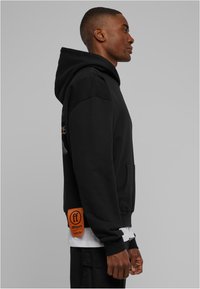 Sudadera negra con capucha, bolsillo canguro y etiqueta de tela naranja. Cuenta con un material ligero y un ajuste relajado con un diseño gráfico.