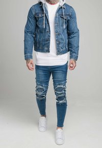 Denimjacka med huva, blå färg, knappstängning fram, tillsammans med trasiga blå skinny jeans och vita sneakers. Har tatueringar på armarna.