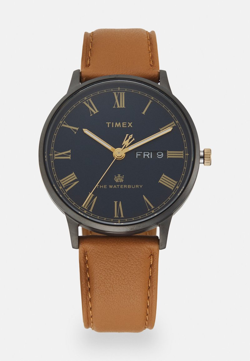 Timex Waterbury kellaga, millel on ümmargune tumesinine kellapealne, kuldsedroomi numbrid ja kuldne kroon; sellel on hebe nahast rihm, millel on nähtavad õmblused.