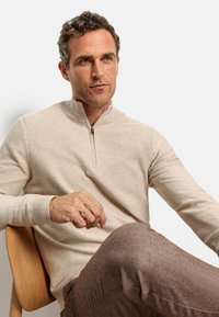 Beige Zip-Pullover mit strukturierten Streifen, langen Ärmeln und gerippten Bündchen, kombiniert mit braunen, schmal zulaufenden Hosen, auf einem Holzstuhl sitzend.