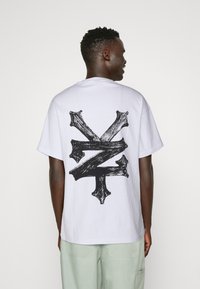 Hvid bomulds T-shirt med et stort sort grafisk design på ryggen, der ligner en abstrakt X-form med teksturede accenter.