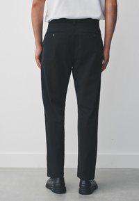 Black cotton trousers with a straight cut, two back pockets, and subtle stitching details. Worn with black shoes.