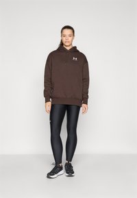 Brun hettegenser med frontlogo, kombinert med svarte leggings og svarte treningssko. Glatt stoff med en avslappet passform.