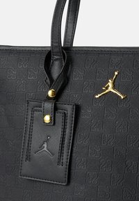 Svart tote bag med preget "23" mønster, teksturert materiale, gull detaljer, og en glidelås etikett med Jumpman-logo. Doble håndtak.