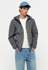 Chaqueta ligera gris con cremallera amarilla y negra, puños elásticos y un parche con logo. Llevada sobre una camisa blanca y combinada con jeans azul claro.