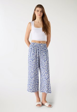 Giovane donna con capelli castani lunghi, in piedi, indossa un top corto senza maniche bianco, pantaloni larghi a fiori blu e bianchi e sandali bianchi a ciabatta.