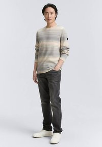 Maglia a maniche lunghe a righe beige, grigie e bianche. Jeans grigi, sneakers bianche. Modello in piedi con le mani nelle tasche su uno sfondo semplice.