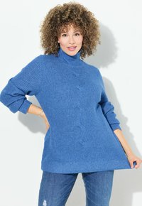 Helles bläuliches, gestricktes Turtleneck-Pullover mit einem Kabelmuster an der Vorderseite, mit fallenden Schultern und gerippten Bündchen. Kombiniert mit blauen Jeans.