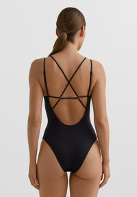 Sort badedragt med tynde, krydsede stropper og et åbent rygdesign, der har en glat tekstur og en figursyet silhuet.