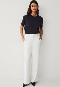 Zwart shirt met korte mouwen gecombineerd met hooggewaide, wijde witte broeken en spitse zwarte hakken; het model heeft haar handen in de zakken.