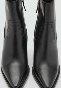 Botas de tobillo de cuero negro con punta afilada, que cuentan con costuras decorativas y un zipper lateral para facilitar su uso. Textura suave.