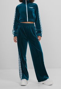 Tyrkysový sametový teplákový komplet s krátkou zipovou bundou s vysokým límcem a bílým vzorem řetězu na rukávech, doplněný odpovídajícími kalhotami.