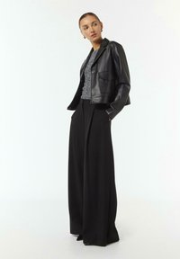 Schwarze Leder-Cropped-Jacke über einem gemusterten Top, kombiniert mit hochtaillierten, weiten schwarzen Hosen. Minimale Hardware und glatte Textur.