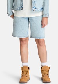 Shorts en denim bleu clair avec une coupe droite, ourlet effiloché, associés à des bottes de travail marron avec des lacets jaunes et des chaussettes blanches à la cheville.