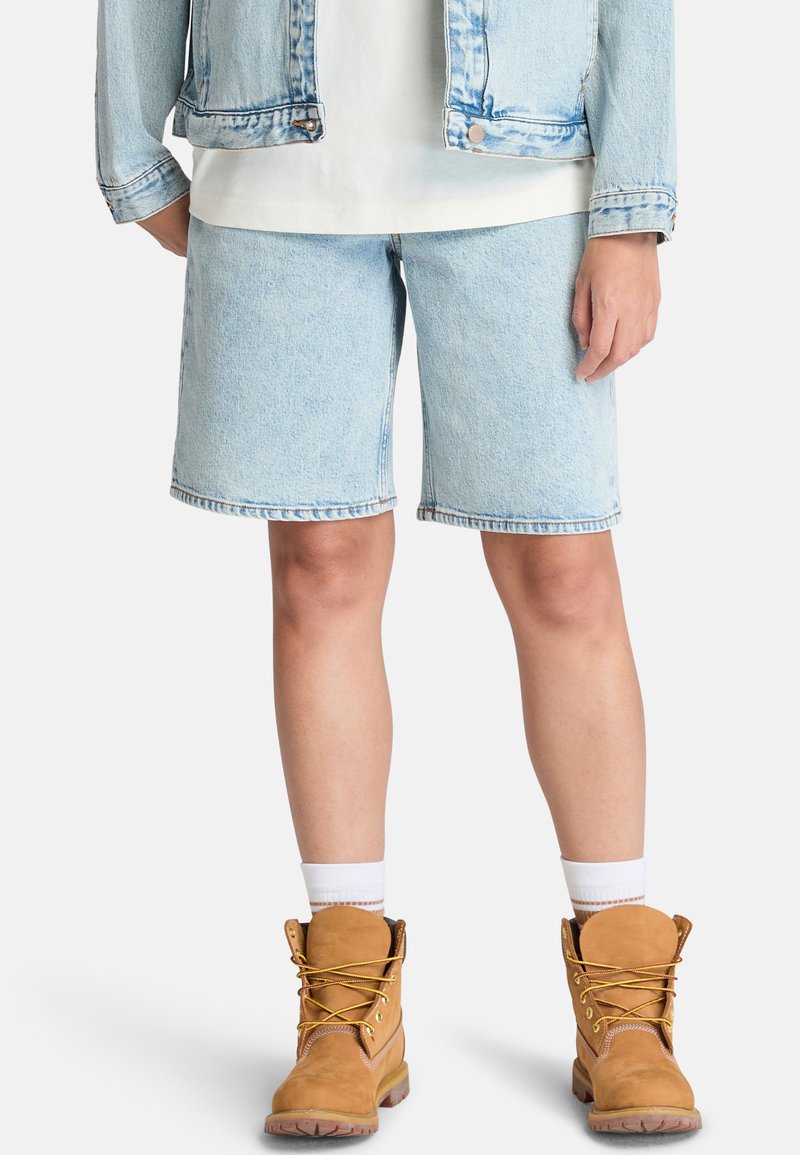Shorts en denim bleu clair avec une coupe droite, ourlet effiloché, associés à des bottes de travail marron avec des lacets jaunes et des chaussettes blanches à la cheville.