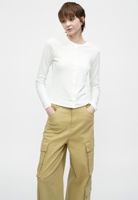 Noor inimene lühikeste pruunide juustega, seljas valge nööbitav pika varrukaga särk ja lahedad beežid cargo-püksid, käed pükste taskutes.