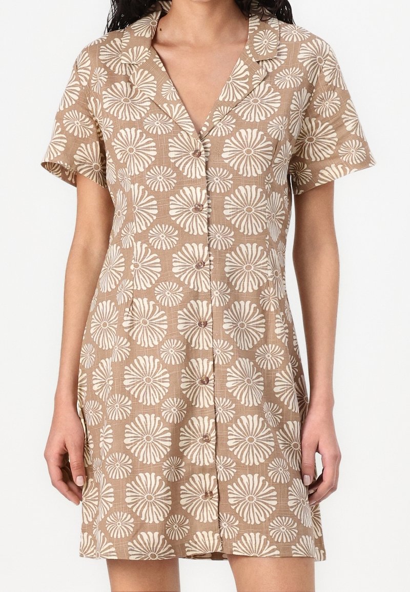 Femme portant une robe beige à manches courtes avec des motifs floraux circulaires blancs et des boutons à l'avant, debout devant un fond uni.