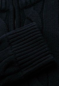 Gros plan d'un tissu tricoté bleu marine foncé avec des détails de poignet côtelés, montrant un motif de tricot en câble texturé et une manche repliée.