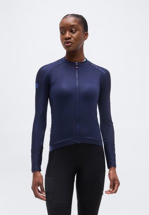 Γυναίκα που φοράει εφαρμοστό μπλε σκούρο μακρυμάνικο ποδηλατικό μπλουζάκι με φερμουάρ μπροστά και μαύρο ποδηλατικό σορτς μπροστά σε απλό φόντο.