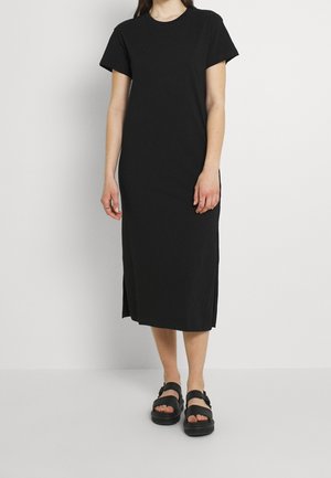 Frau trägt ein schwarzes Midikleid mit kurzen Ärmeln, seitlichen Schlitzen und schwarzen Sandalen, steht vor einem schlichten hellen Hintergrund.