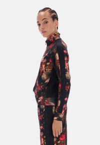 Mladá osoba s vrkočmi má na sebe bundu s kvetinovým vzorom a zodpovedajúce nohavice s čiernymi pruhmi, stojí bokom na bielom pozadí.