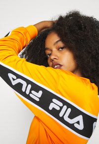 Giacca sportiva arancione con dettagli a banda con logo nero e bianco sulla manica. Realizzata in materiale morbido e con una vestibilità rilassata.