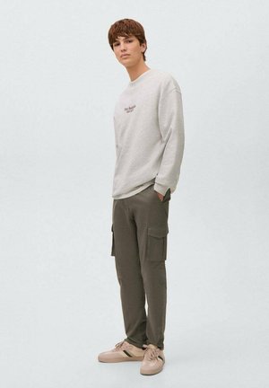 Giovane uomo in piedi con le mani nelle tasche, che indossa una felpa grigio chiaro con scritte e pantaloni cargo scuri con sneakers beige.