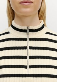 Gestreifter, cremefarbener und schwarzer hochgeschlossener Pullover mit Rippstrick, kurzem Frontreißverschluss und weicher Textur.