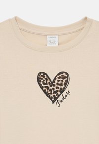 Beżowy t-shirt z okrągłym dekoltem, na którym znajduje się serce z czarnym konturem wypełnione wzorem w panterkę oraz napis "J'adore" w kursywie poniżej.