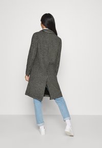 Abrigo largo de patrón de espiga en blanco y negro con un ajuste entallado, abertura en la espalda, llevado sobre jeans azules claros y zapatillas blancas.