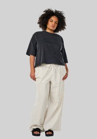 Sort oversized T-shirt med tekst, parret med løse hvide stribede bukser. Sandaler har et design med sorte stropper. Neutral baggrund.
