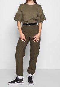 Femme portant un haut à manches volantes olive, un pantalon cargo taille haute avec une ceinture noire, des chaussettes blanches et des baskets noir et blanc.