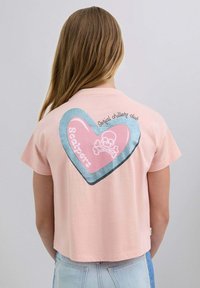 Camiseta rosa corta con un gran gráfico de corazón que presenta una calavera y tibias cruzadas, el texto dice "Club de los que chillan en serie" en una fuente juguetona.