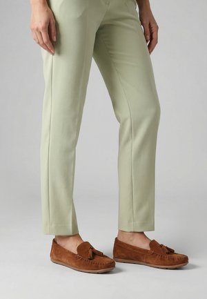 Persoon draagt lichtgroene taps toelopende broek en bruine suède loafers met kwastjes, staand op een effen grijze ondergrond.