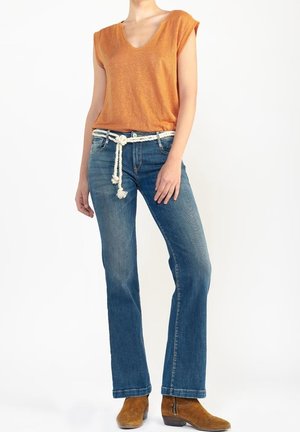 Kvinde iført orange kortærmet V-hals shirt, blå jeans med et hvidt reb bælte og brune ruskind ankelstøvler, stående mod en hvid baggrund.