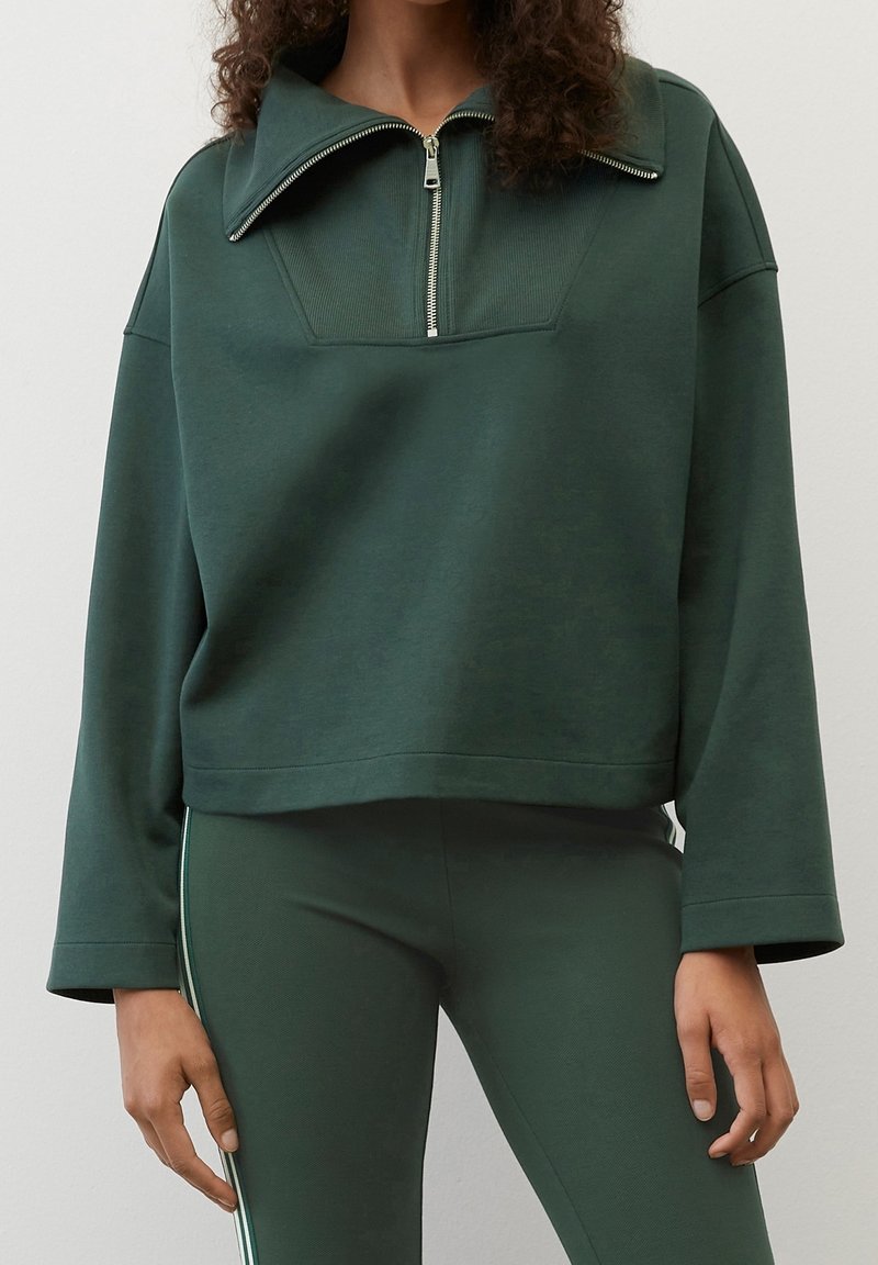 Sweat-shirt cropped vert foncé avec un col haut et une fermeture éclair. Présente des détails côtelés et des manches larges. Associé à des leggings assortis.
