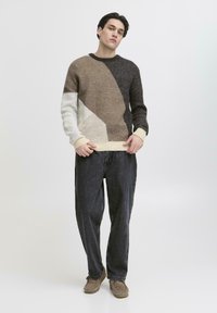 Un tânăr stând cu mâinile lângă tivul puloverului, îmbrăcat cu un pulover multicolor în nuanțe de marron, negru și bej, blugi largi negri și pantofi maro.