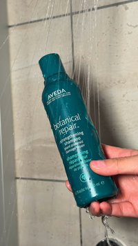 Eine Hand hält eine Flasche des Aveda Botanical Repair stärkenden Shampoos unter fließendem Duschwasser, im Hintergrund beige geflieste Wände.