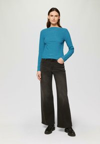 Haut à manches longues en ribbed teal associé à un jean noir à jambes larges et des bottes noires à lacets, présentant une texture subtile sur le tissu.