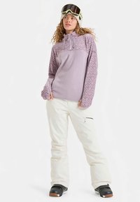 Lichtpaarse trui met gestructureerde mouwen, gecombineerd met witte skibroek en zwarte snowboardlaarzen. Voorzien van een ritssluiting bij de hals en schreefloze accenten.