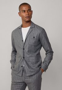 Giacca slim-fit grigia a righe sottili realizzata in tessuto texture, con colletto a spacco, due tasche frontali e chiusura a bottoni. Indossata sopra una camicia bianca.
